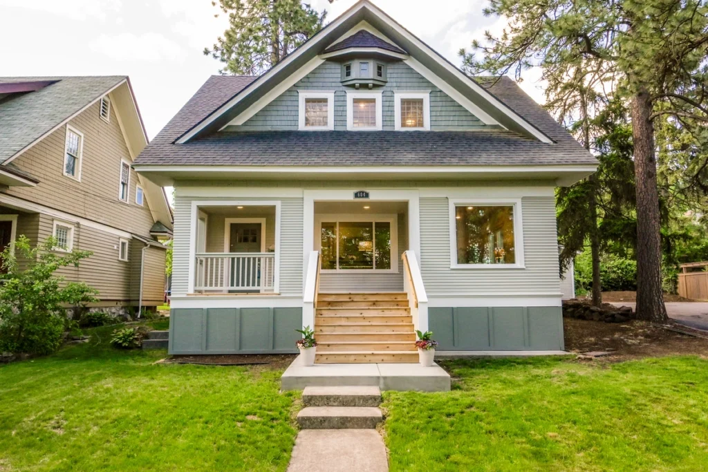 A two-story house with a gabled roof adorned with architectural shingles boasts a covered front porch, central staircase, and manicured lawn. Large windows on both floors let in light, while surrounding trees complete the picturesque scene.
