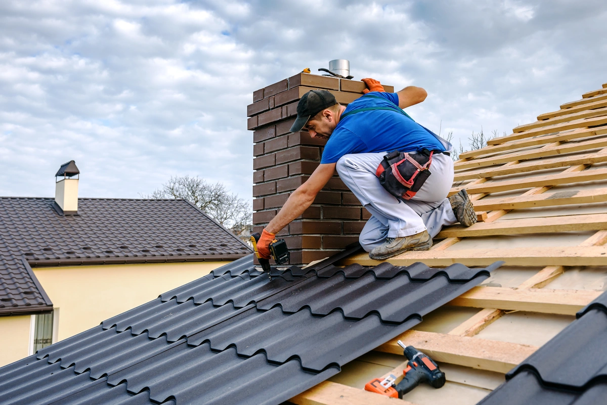 A construction worker in a blue shirt and white pants demonstrates how to install metal roofing by fitting black sheets on a sloped roof beside a brick chimney. With a drill in hand and other tools scattered nearby, he works efficiently under the cloudy sky.