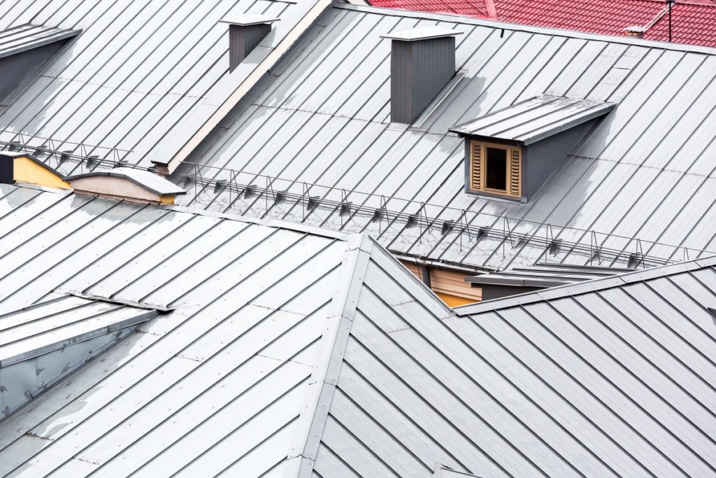 Aerial view of intersecting metal roofs with triangular and rectangular dormers showcases a blend of grey and vibrant red shingles, forming a geometric pattern. Despite the intricate design, the efficient layout reflects practical metal roof cost benefits, enhanced by small ladders and railings.