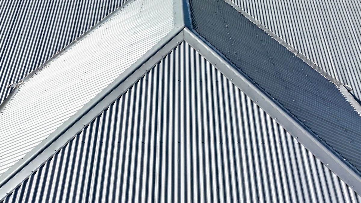 Close-up of corrugated metal roofing with a symmetrical triangular peak. The texture of the grooves creates a geometric pattern, with shadows enhancing its three-dimensional appearance. The shiny surface reflects light beautifully.