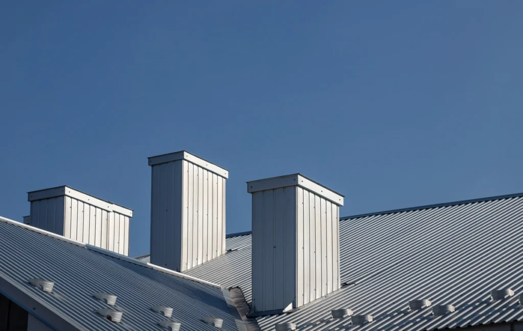 A rooftop showcases three white metal chimneys against a clear blue sky, with the corrugated metal roofing beautifully reflecting the sunlight.