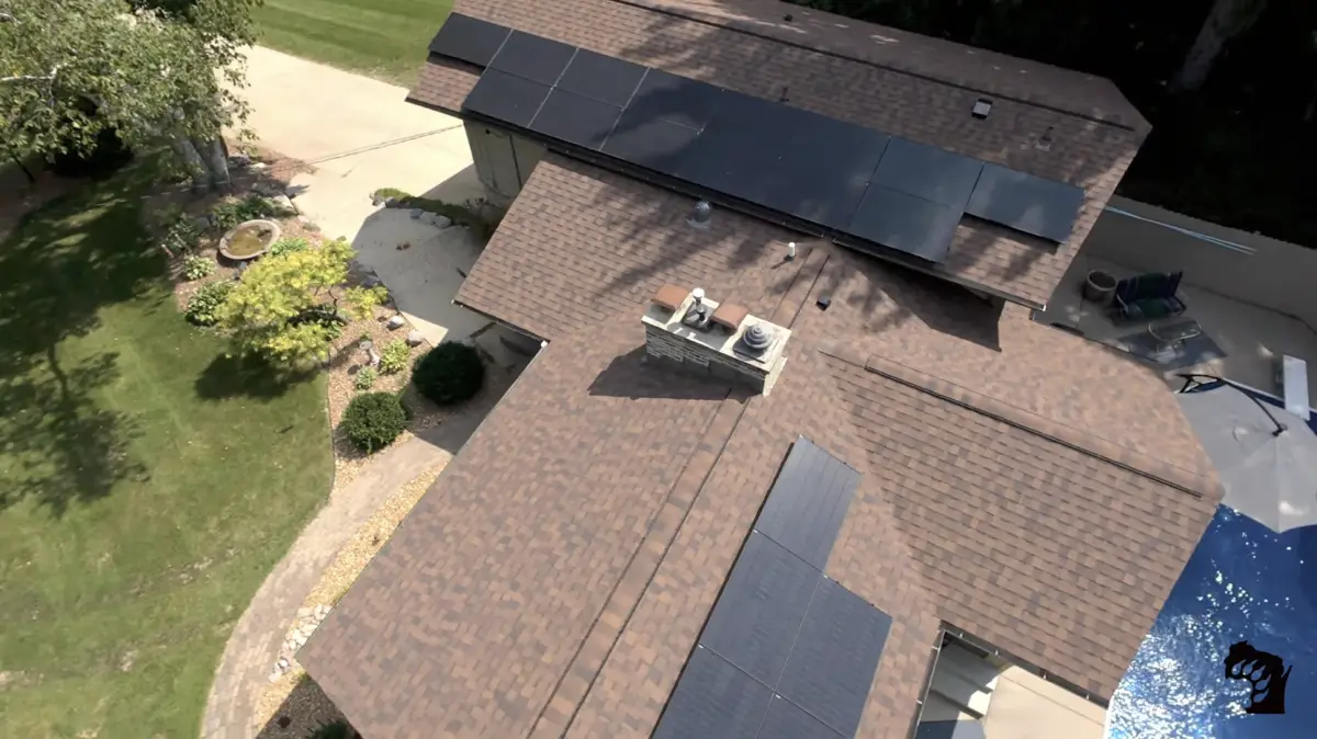 Aerial view of a house with multiple solar panels installed on its brown shingle roof, surrounded by green grass, a landscaped garden, and a driveway.