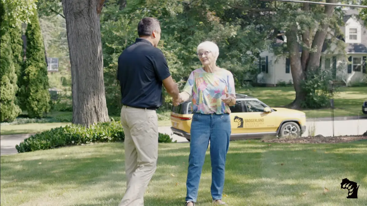 A man and an older woman shake hands on a grassy lawn in a residential neighborhood, with a yellow car labeled "ROOFING" parked on the street in the background.