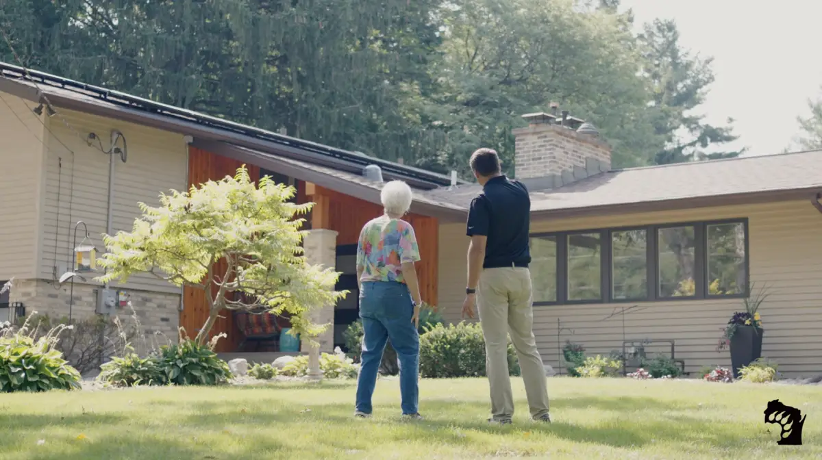 An elderly person and a younger adult stand on a grassy lawn, facing a house with a sloped roof and looking up, possibly discussing something related to the home exterior. Trees and plants surround the property.