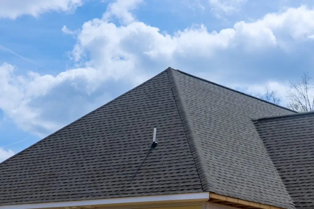 A close-up view of a house's gray shingled roof in Lower Burrell, PA, set against a bright blue sky filled with fluffy white clouds. A small antenna is visible on the roof's peak.