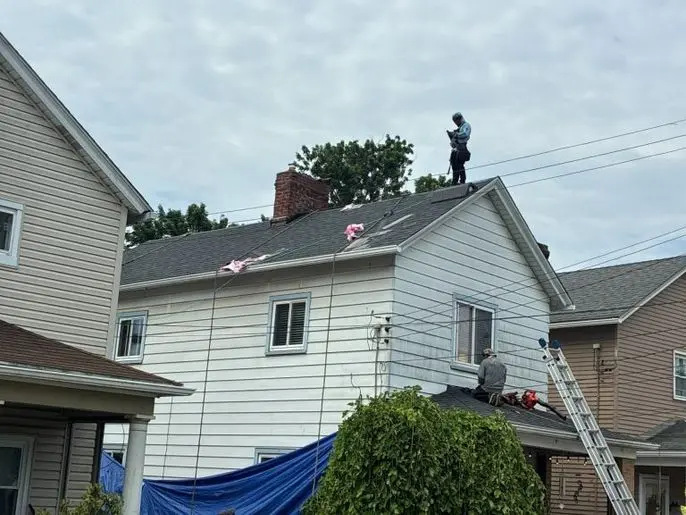 Two workers repair the roof of a white house; one stands on the roof holding tools, while the other kneels near a ladder. Wires, debris, and a blue tarp are visible. The sky is cloudy.