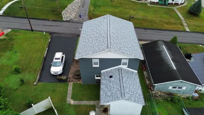 Aerial view of a suburban area featuring a two-story gray house with a gray roof. A white car is parked in the driveway. The surrounding area includes green lawns and intersecting roads. Another house with a dark roof is adjacent.