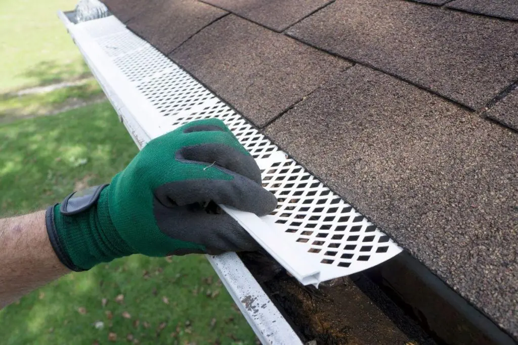 A person wearing a work glove installing a white metal gutter guard over a roof gutter.