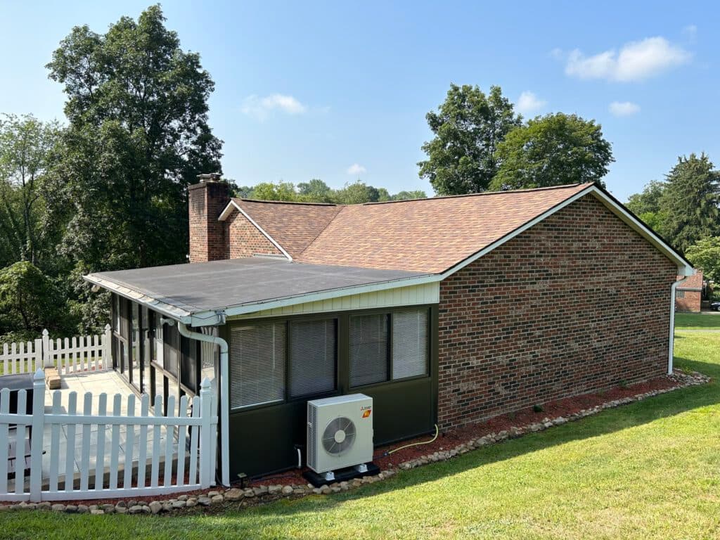 A brick house with a fenced patio and a sloped roof sits on a grassy lawn. An outdoor air conditioning unit is mounted by the patio. Tall trees are in the background under a clear blue sky.