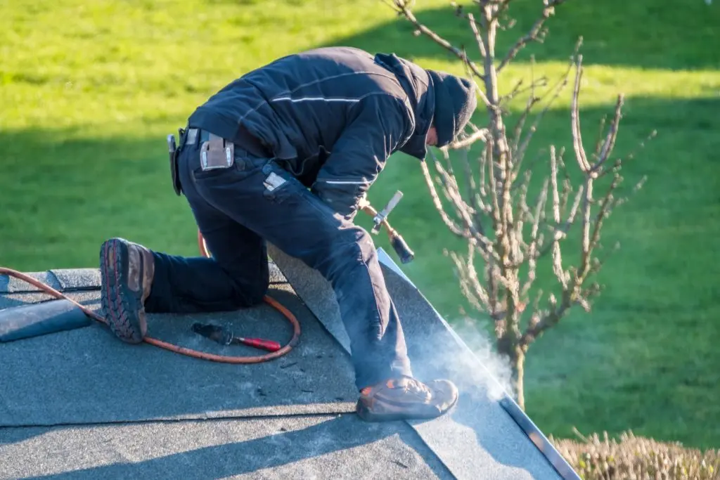 A worker in dark clothing kneels on a roof, using a torch and tools to install roofing material, with smoke rising and green grass in the background.