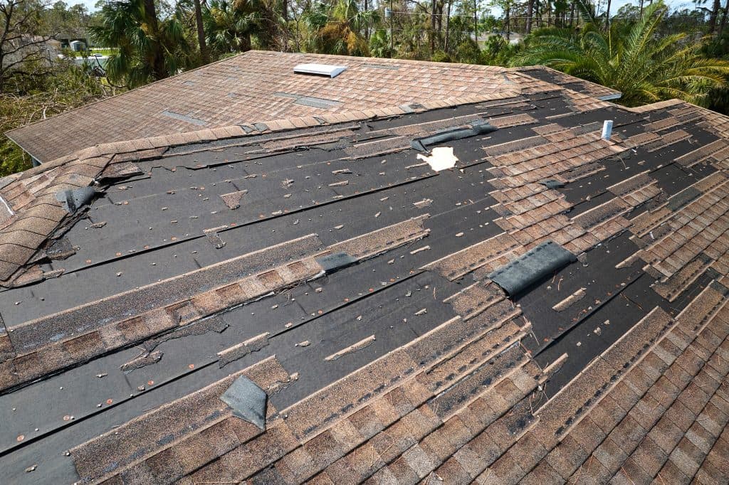 Damaged shingled roof with missing and displaced shingles, exposing the black underlayment. Surrounding trees and another section of the house roof are visible in the background.