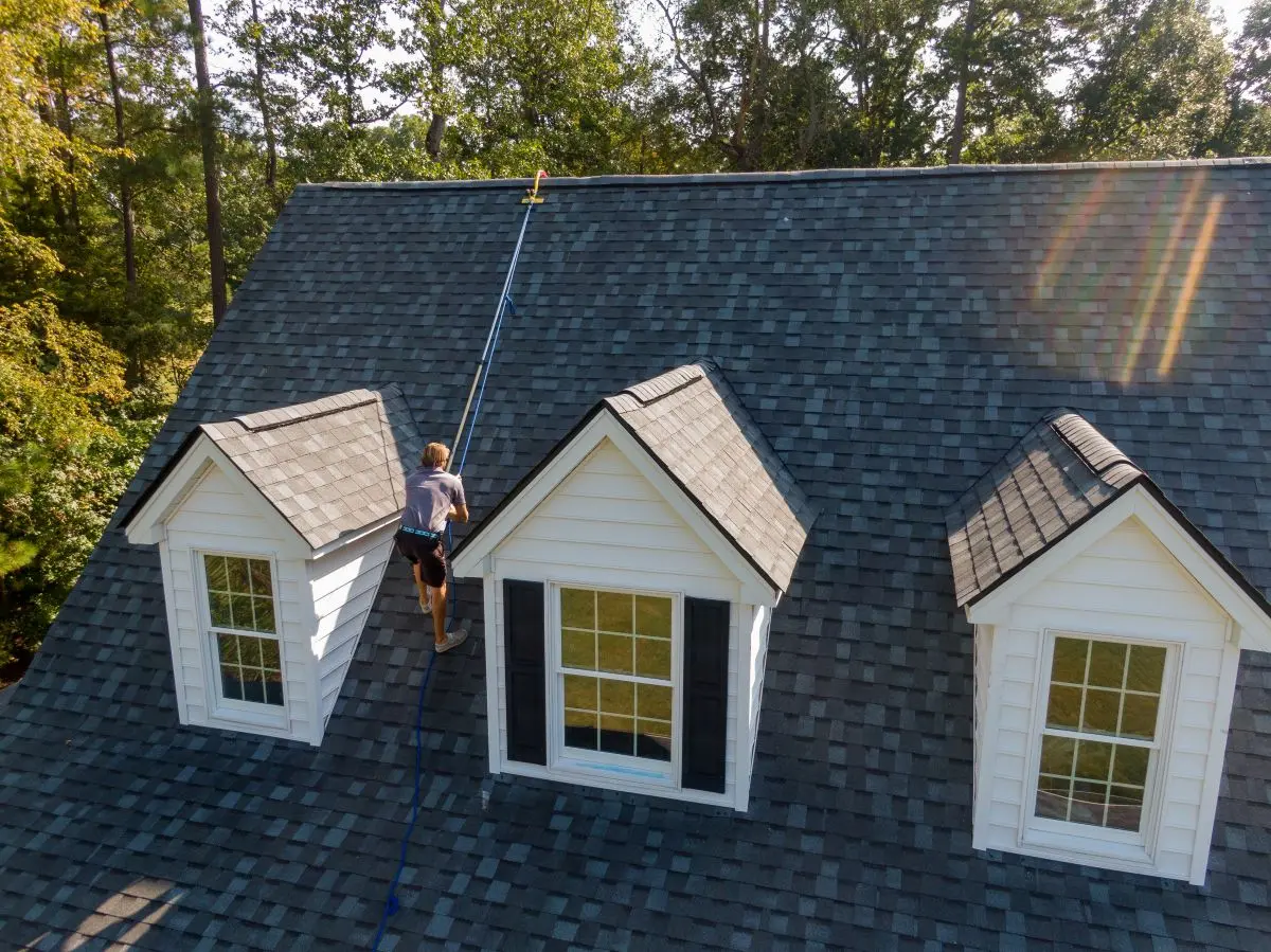 A person cleans a steep, gray-shingled house roof using a long-handled tool on a sunny day, surrounded by trees.