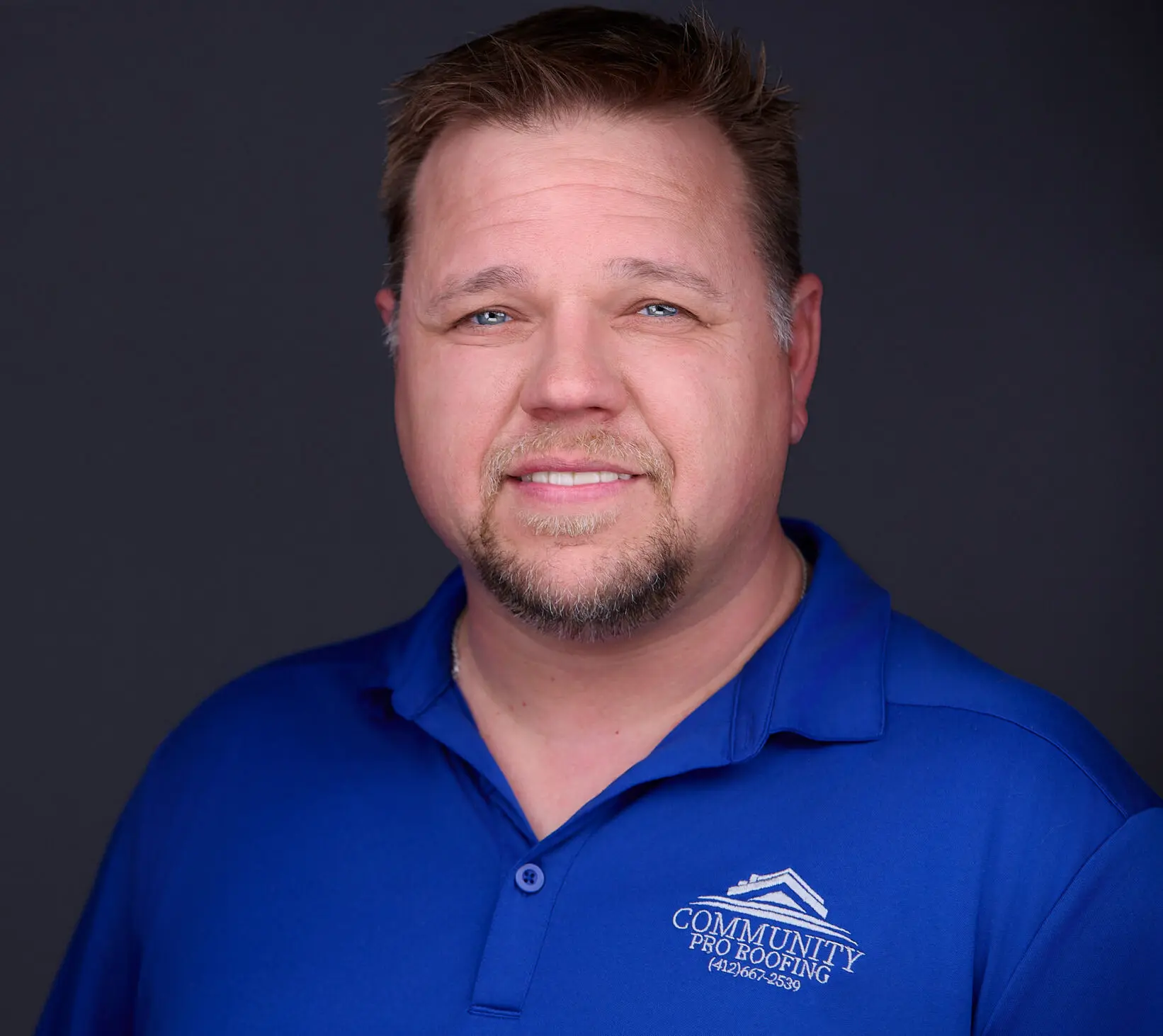 Man in a blue polo shirt with a company logo standing against a dark background, smiling at the camera.