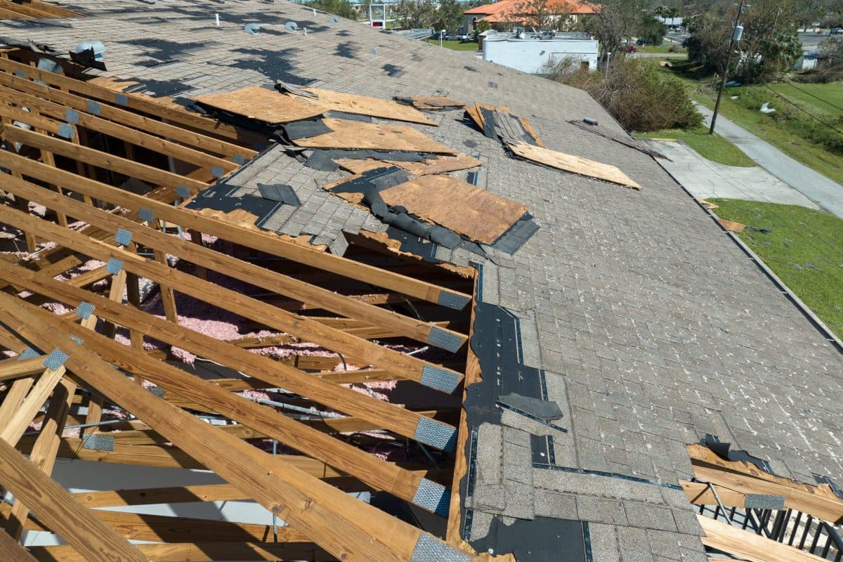 storm damage roof repair A roof with missing shingles and exposed wooden beams, showing severe storm damage. Pieces of plywood are scattered across the damaged area, revealing the interior of the house below.