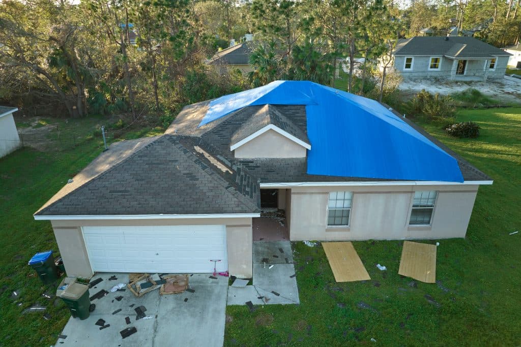 A single-story house with a blue tarp covering part of its roof, debris scattered on the driveway and lawn, suggesting recent roof damage.