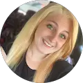 A person with long, straight blonde hair smiles at the camera. They are wearing a light-colored top, and the background is slightly blurred, showing a mix of colors.
