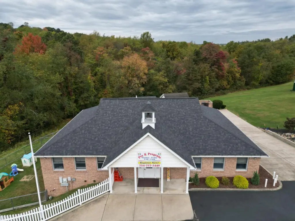A single-story brick building with a dark gray roof, surrounded by a large lawn and leafy autumn trees. A playground set is visible to the left. There's a long driveway, and a sign displayed on the building's front. The sky is overcast.