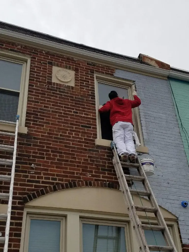 A house painter in a red hoodie and white work pants stands on a ladder painting the upper front wall and window trim of a two-story brick rowhouse.