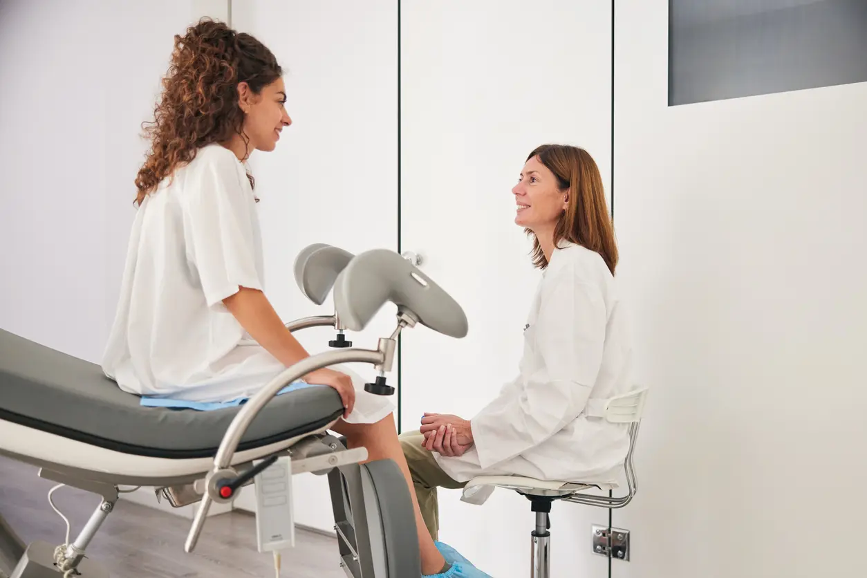 The young woman, dressed in a hospital gown, sits on the stretcher as the mature doctor in a white lab coat sits on a stool, and they engage in discussion, looking at each other.