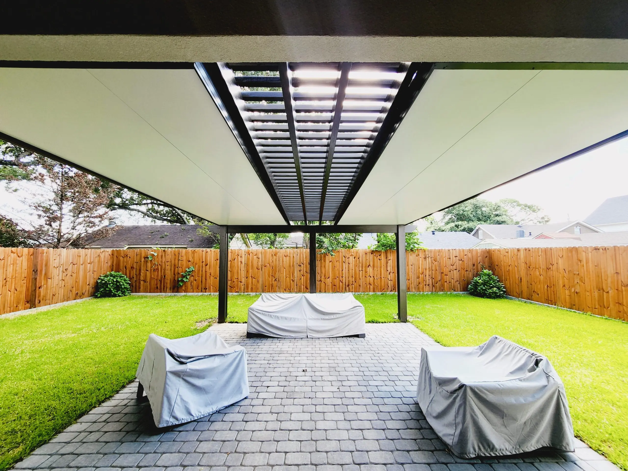 Indoor view of a luxurious outdoor patio with a 4-inch insulated aluminum roof paneled cover and elegant pergola, featuring covered furniture, green grass, and wooden fencing, designed for comfortable outdoor living in Metairie, Louisiana.