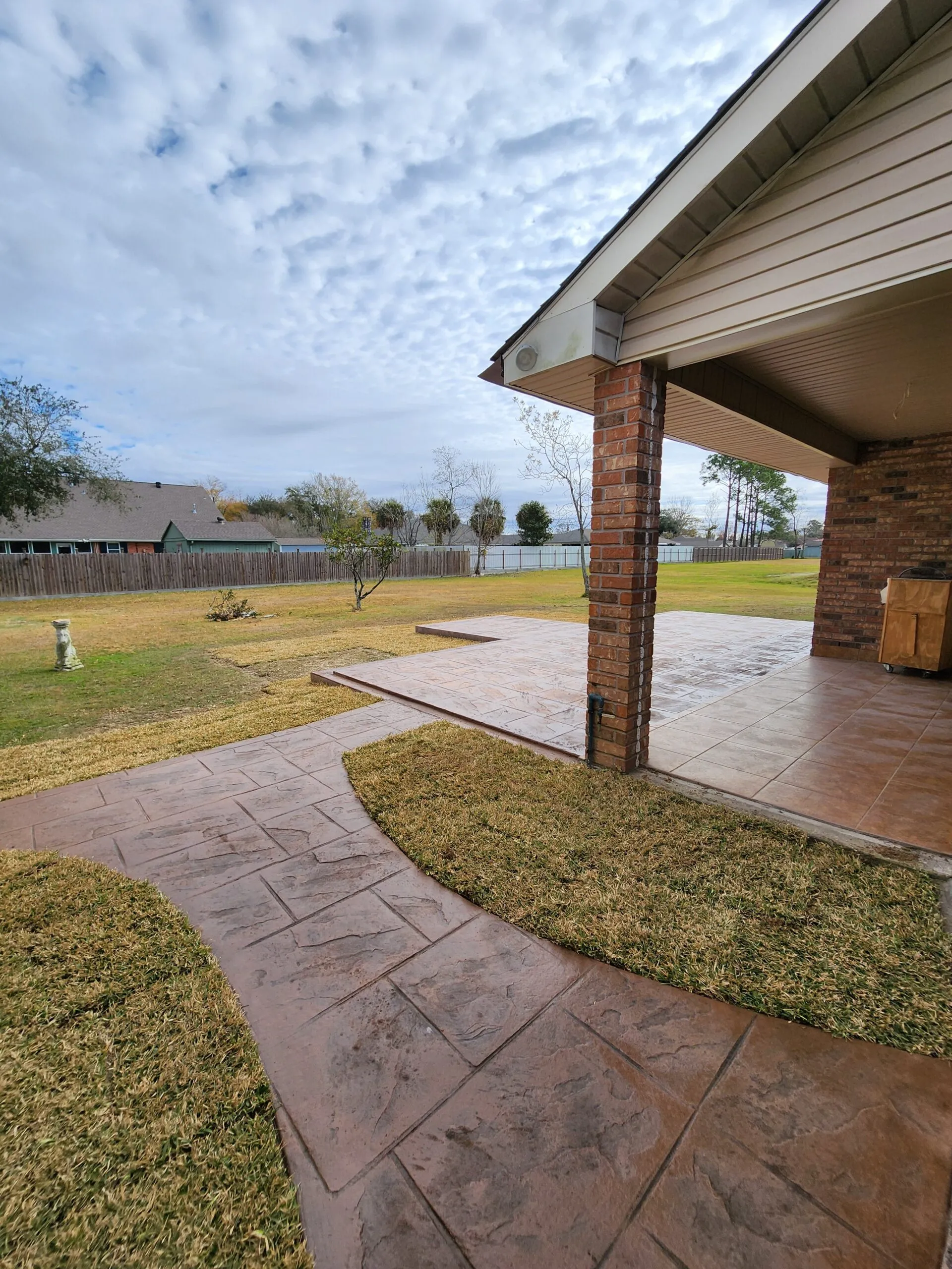 Concrete patio and walkway with decorative stamped finish, surrounded by green grass and a clear sky, showcasing outdoor space for home improvement projects.