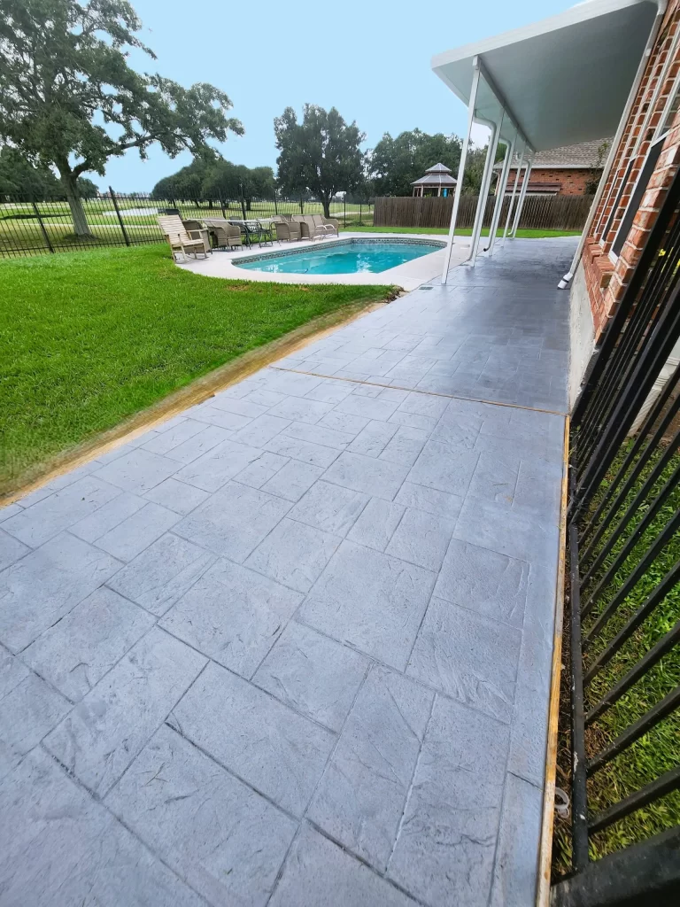 Concrete patio with stamped finish adjacent to a swimming pool, surrounded by green grass and a fenced yard, showcasing outdoor living space enhancements by Capital Improvement.