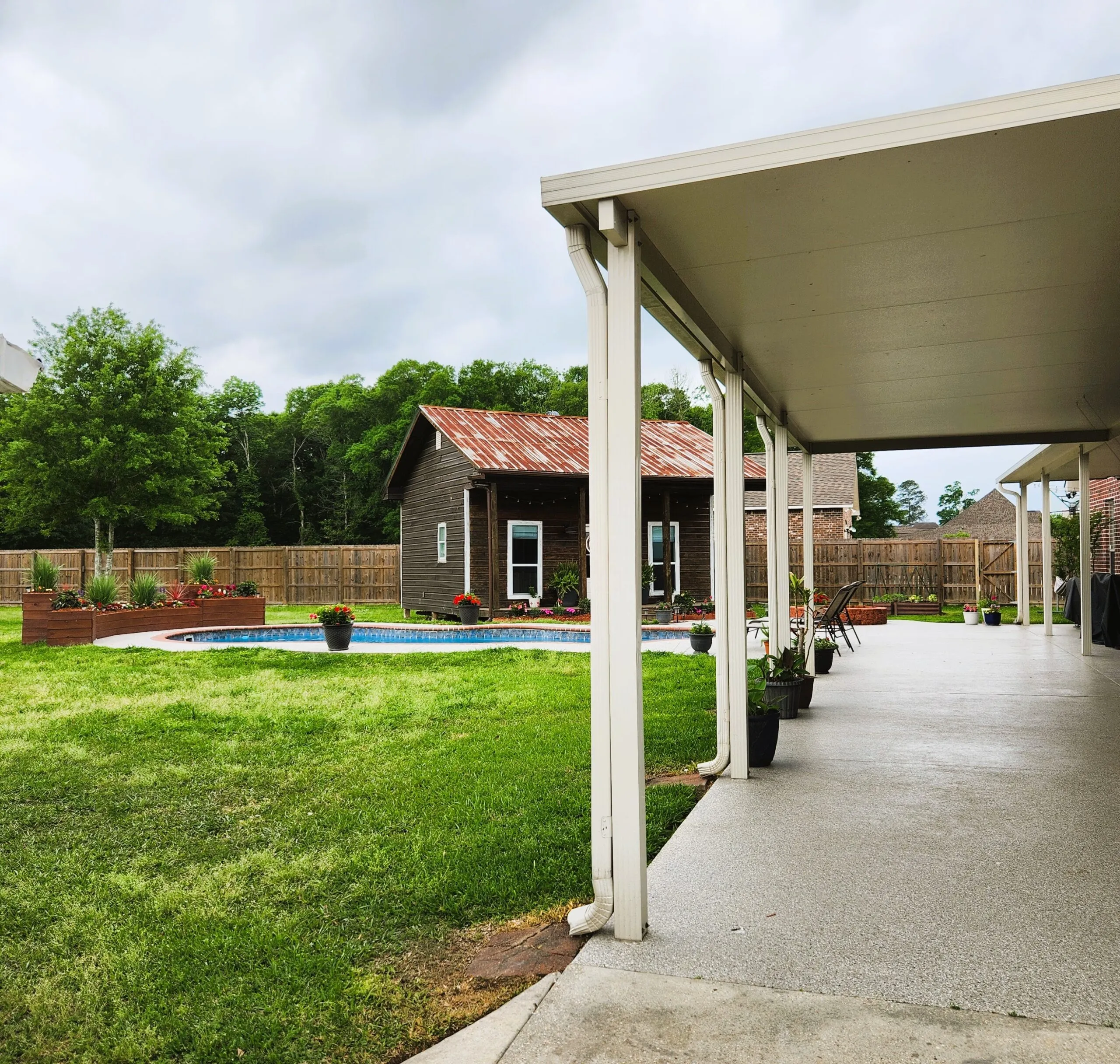Patio cover providing shade over a concrete patio, overlooking a landscaped yard with a pool and rustic wooden shed, showcasing outdoor living space enhancements.