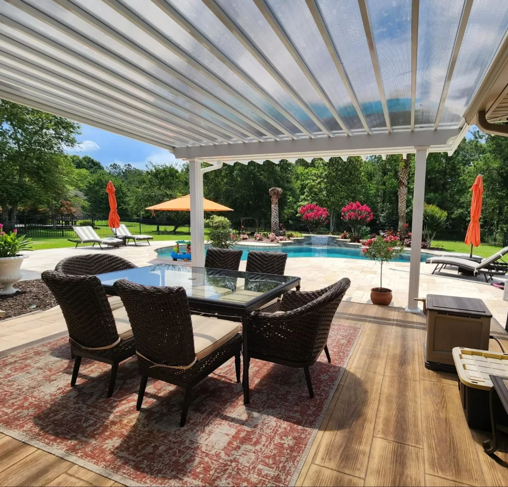 Outdoor patio featuring a glass-top dining table and wicker chairs under a modern patio cover, with a swimming pool and vibrant landscaping in the background, highlighting 2025 patio cover trends for enhancing outdoor living spaces.