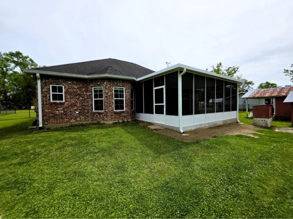 Screen room addition to a home in Metairie, LA, featuring large windows and a white frame, surrounded by a green lawn and trees, enhancing outdoor living space.