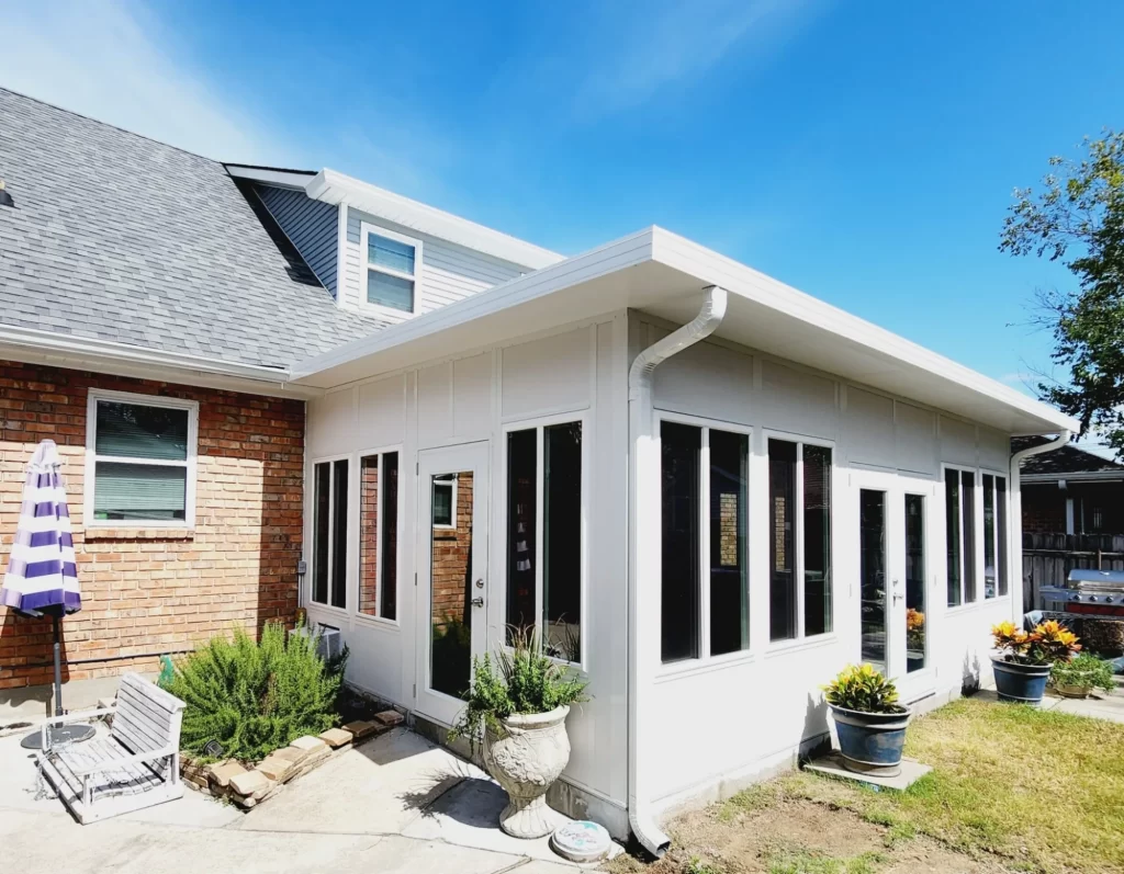 Custom sunroom addition on a home, designed for Louisiana weather, featuring large windows, a white exterior, and adjacent outdoor seating with potted plants.