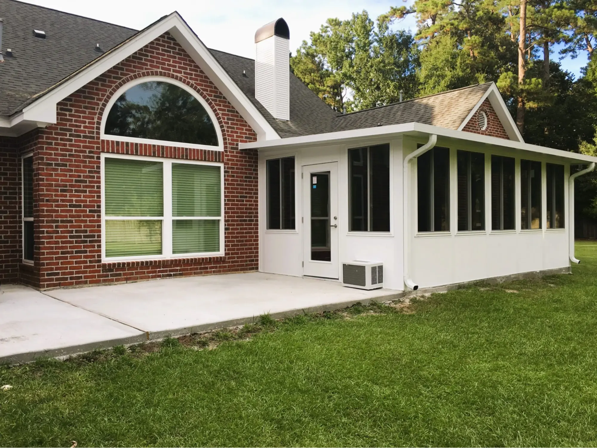 Sunroom with large windows and brick exterior, featuring a white frame and air conditioning unit, set in a green lawn, emphasizing outdoor living space and potential hurricane readiness.