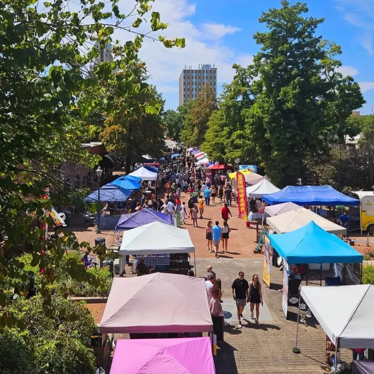Elevated wide view of a downtown pedestrian street market with rows of colorful vendor tents and a crowd of people walking, city high-rise in background, sunny day, Augusta, GA