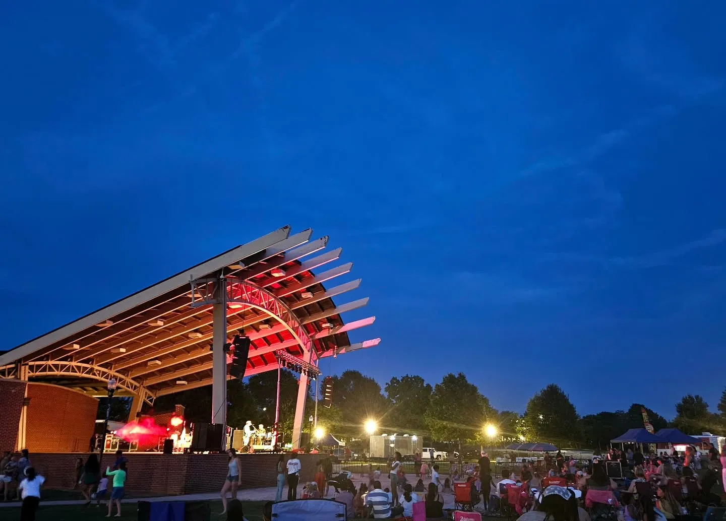 Columbia County Amphitheater, Evans, GA: outdoor amphitheater concert at dusk, band performing on covered stage with red uplighting, crowd seated on lawn in front, blue twilight sky