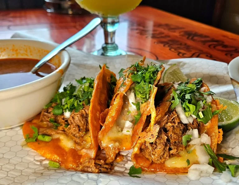 El Kiosco, Grovetown, GA: three birria-style quesabirria tacos with melted cheese, shredded beef, cilantro and onion garnish, lime wedges, served with a bowl of consomme dipping broth and a mango-colored drink in backgro