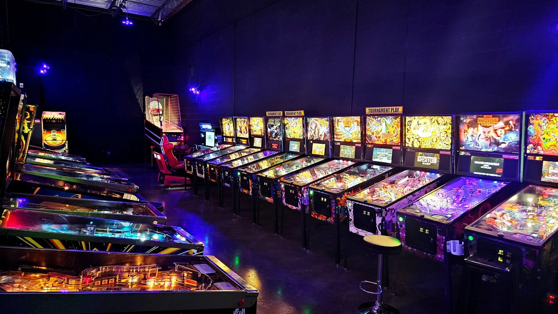 Retro City Arcade, Evans, GA: dark arcade interior lined with rows of lit pinball machines under purple mood lighting, a bar stool in foreground
