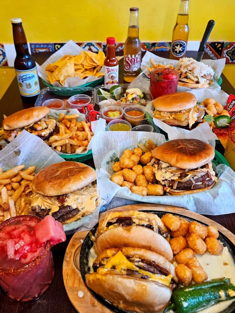 Smash Lab Burgers, Augusta, GA: table spread of multiple smash burgers with melted cheese on baskets of tater tots and fries, tortilla chips, hot sauce, Mexican beer bottles (Corona, Sol), a michelada-style drink with wa