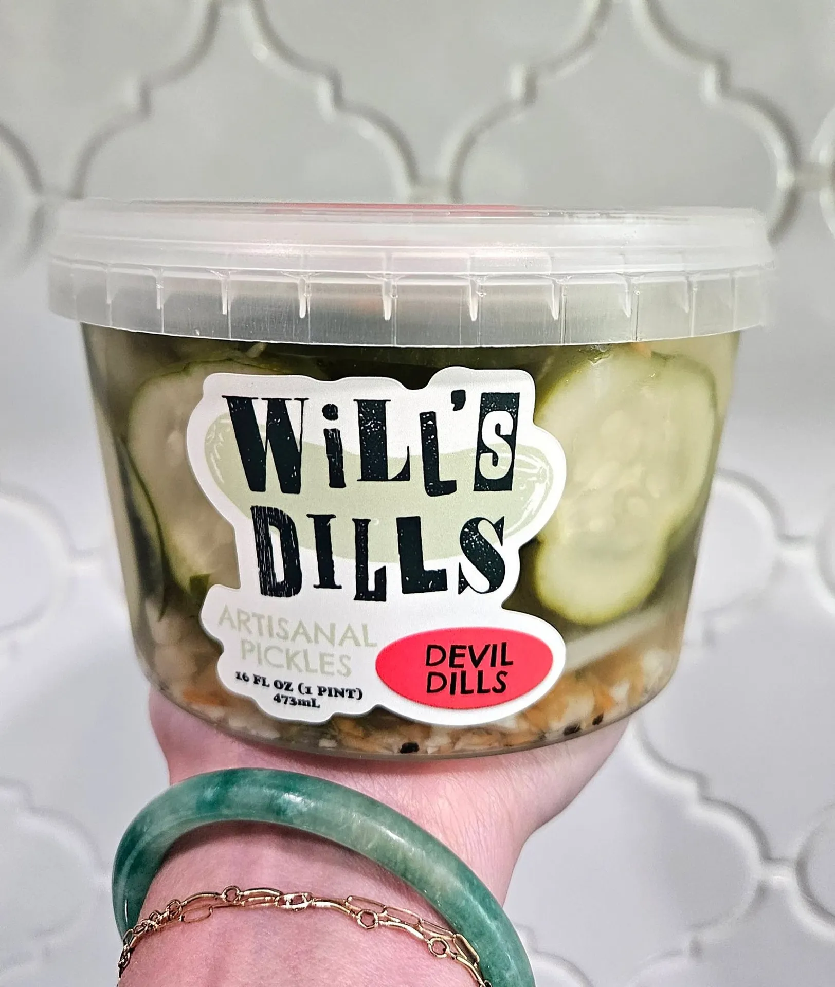Will's Dills, Augusta, GA: hand holding a clear plastic pint container of pickle chips labeled Will's Dills Artisanal Pickles, Devil Dills flavor, tiled background