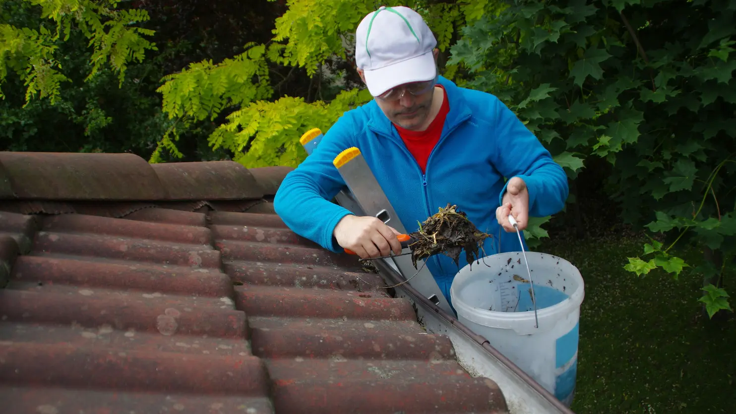 Person cleaning gutters on a roof, wearing a blue jacket and cap, holding debris with a tool, with a bucket nearby, surrounded by green foliage.