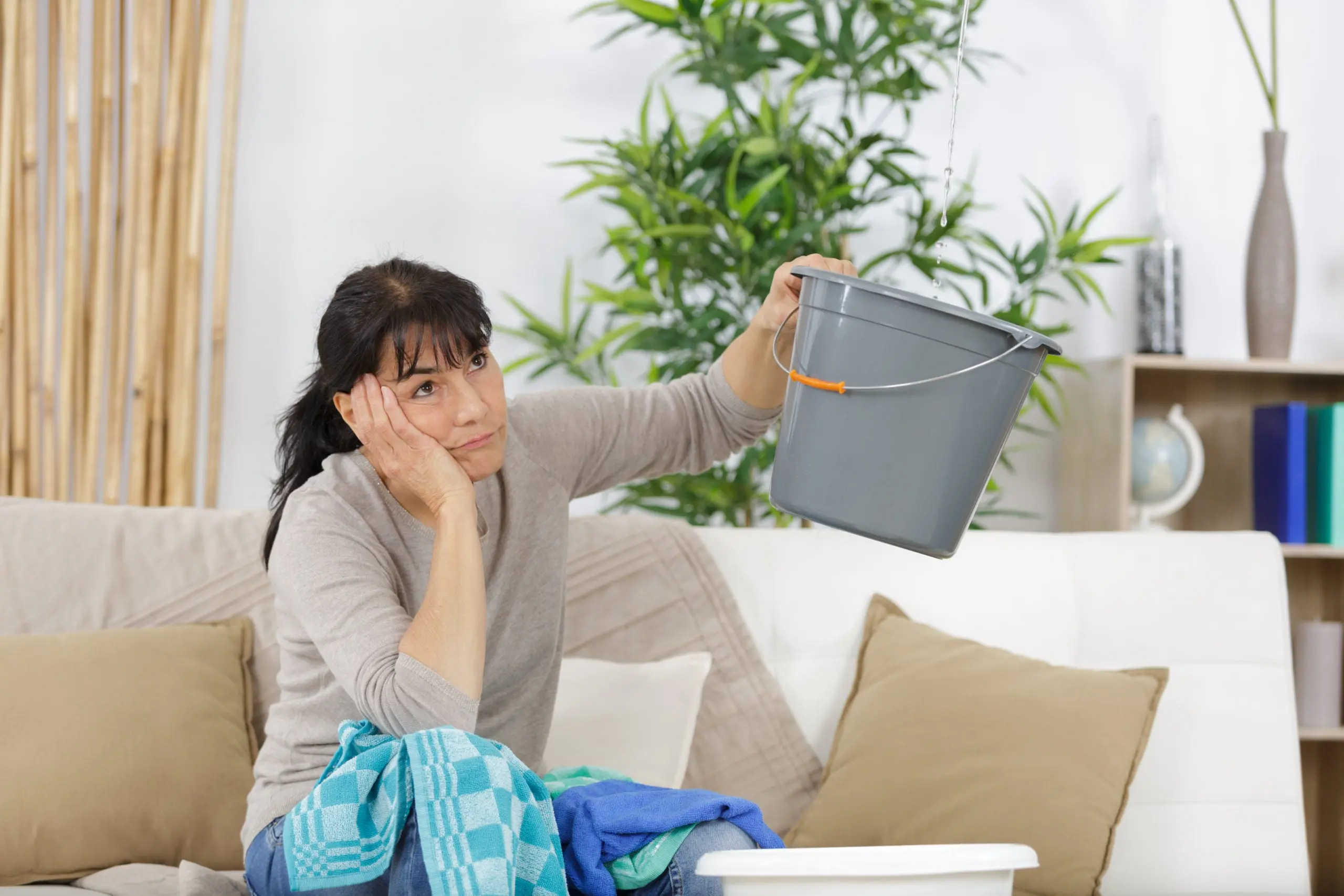 Woman sitting on a couch holding a bucket to catch water dripping from the ceiling, with a towel draped over her lap, expressing concern about a roof leak.