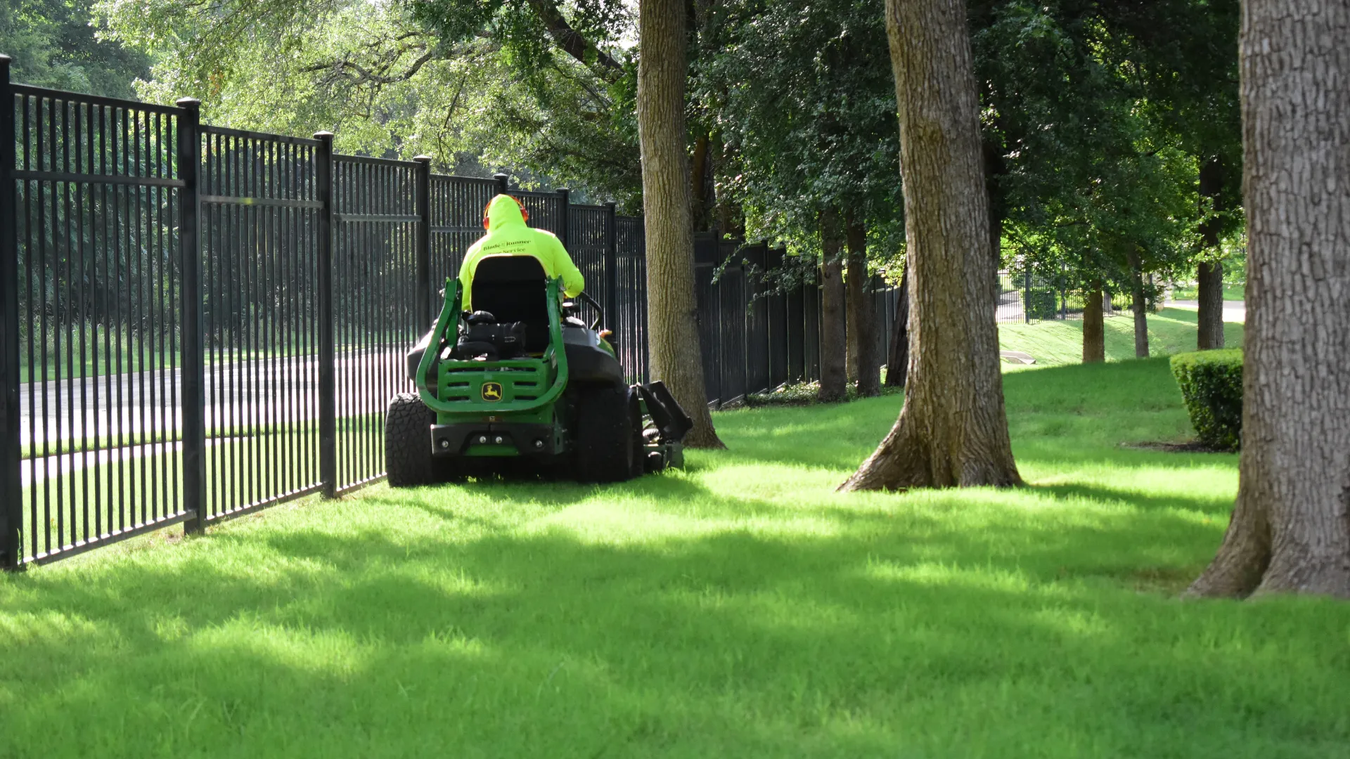 Man on riding lawn mower cutting grass along a rod iron fence