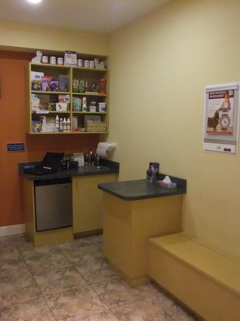 Bright veterinary consultation and prep room featuring light wood cabinetry, a mini-fridge, and stocked wall shelving.