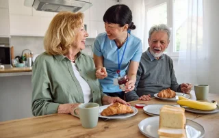 Caregiver assisting older adults with breakfast in a home-style assisted living kitchen.