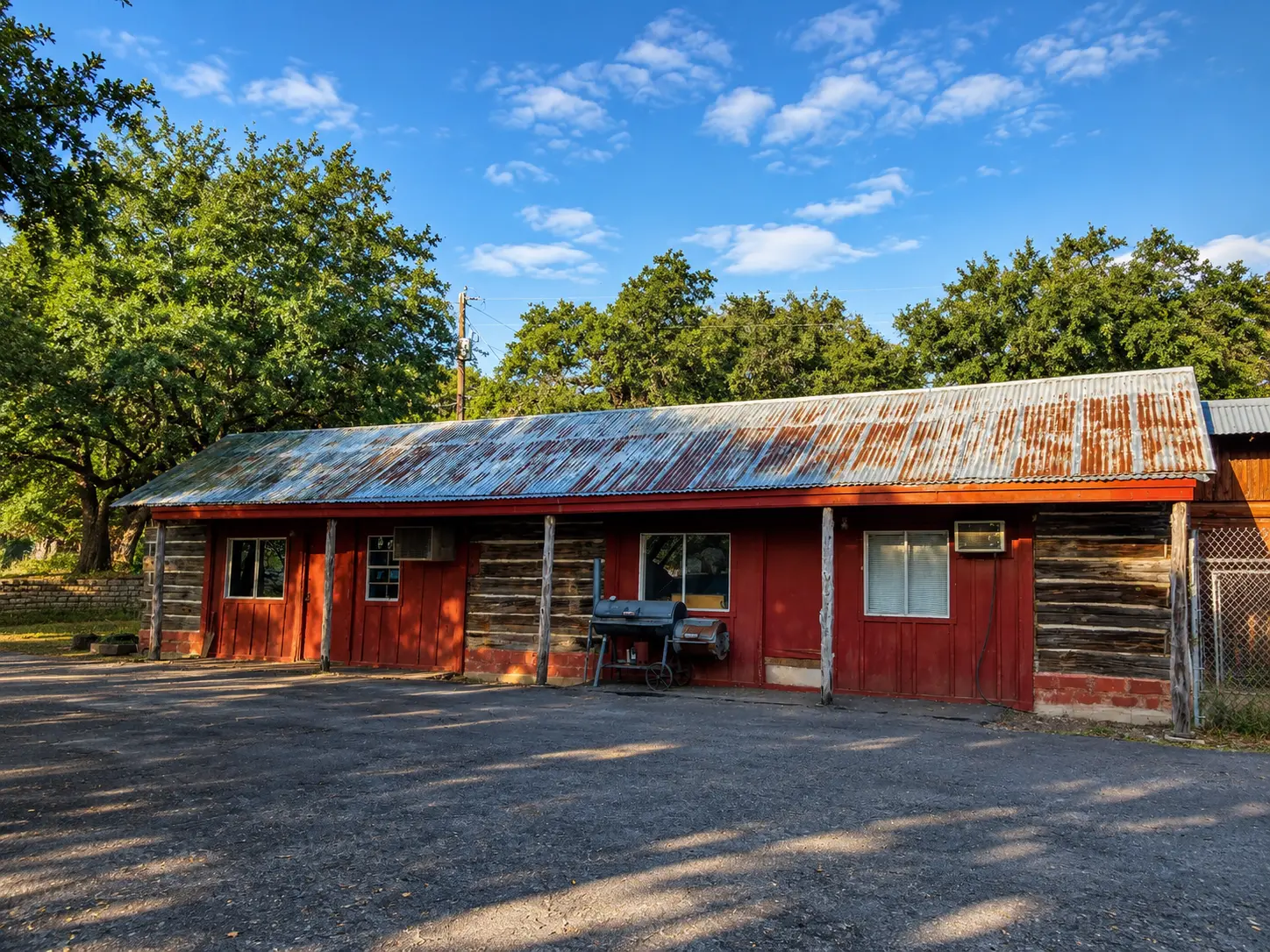 Rustic one-story red wooden building with a corrugated, rusting metal roof; a barbecue grill sits outside in a sunny yard with trees in the background.