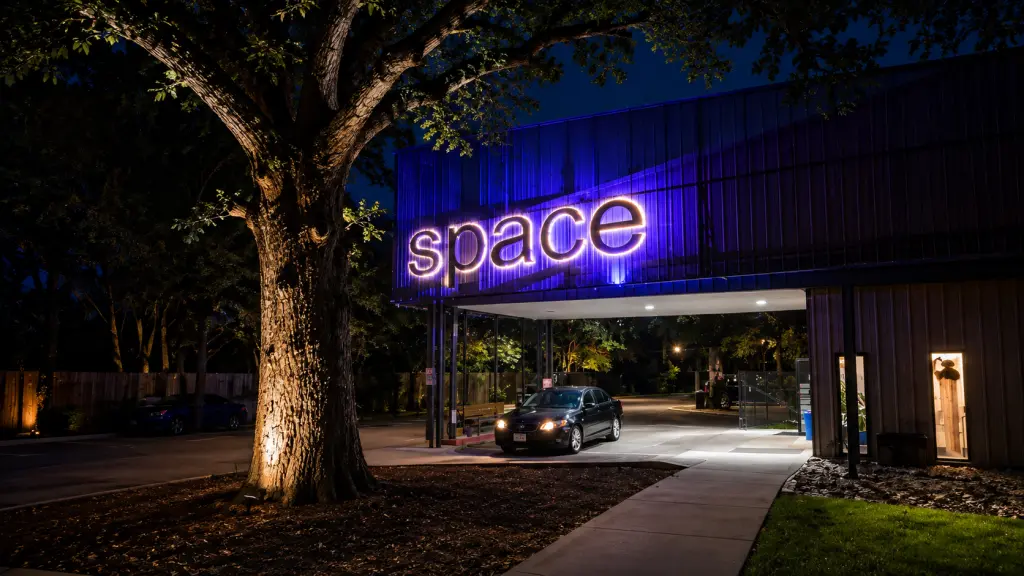 Nighttime view of a commercial building featuring a glowing illuminated "space" sign, architectural blue accent lighting, and an illuminated entrance driveway framed by a mature tree.