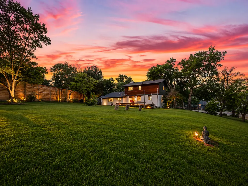 Two-story house with white walls and dark upper level sits in a spacious yard at sunset, illuminated by garden lights in the yard.
