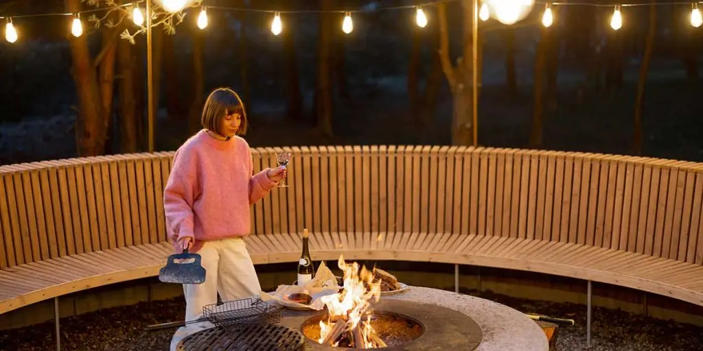 A woman in a pink sweater holds a wine glass near a glowing fire pit surrounded by curved wooden seating.