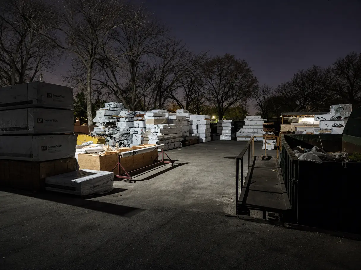 Nighttime view of an outdoor commercial storage yard illuminated by security lighting, featuring stacked building materials and a large dumpster.