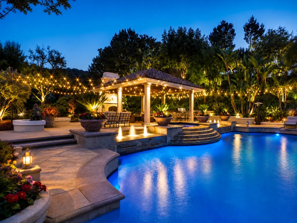 A luxurious pool and patio feature warm string lights, illuminated fountains, tropical landscaping, and a covered outdoor pavilion at dusk.
