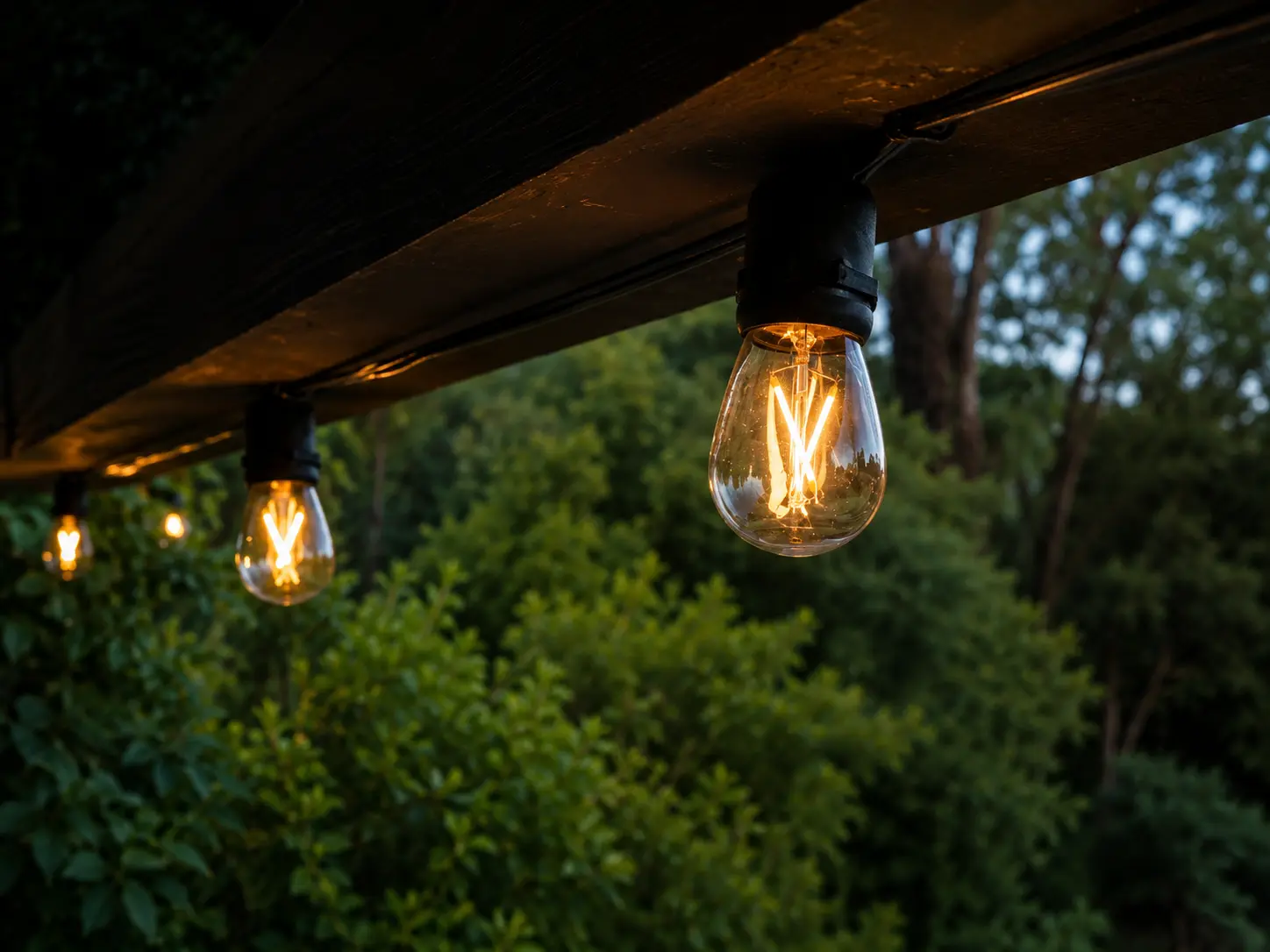 Vintage-style Edison bulbs glow along a wooden pergola structure, creating a warm and inviting outdoor setting at dusk.