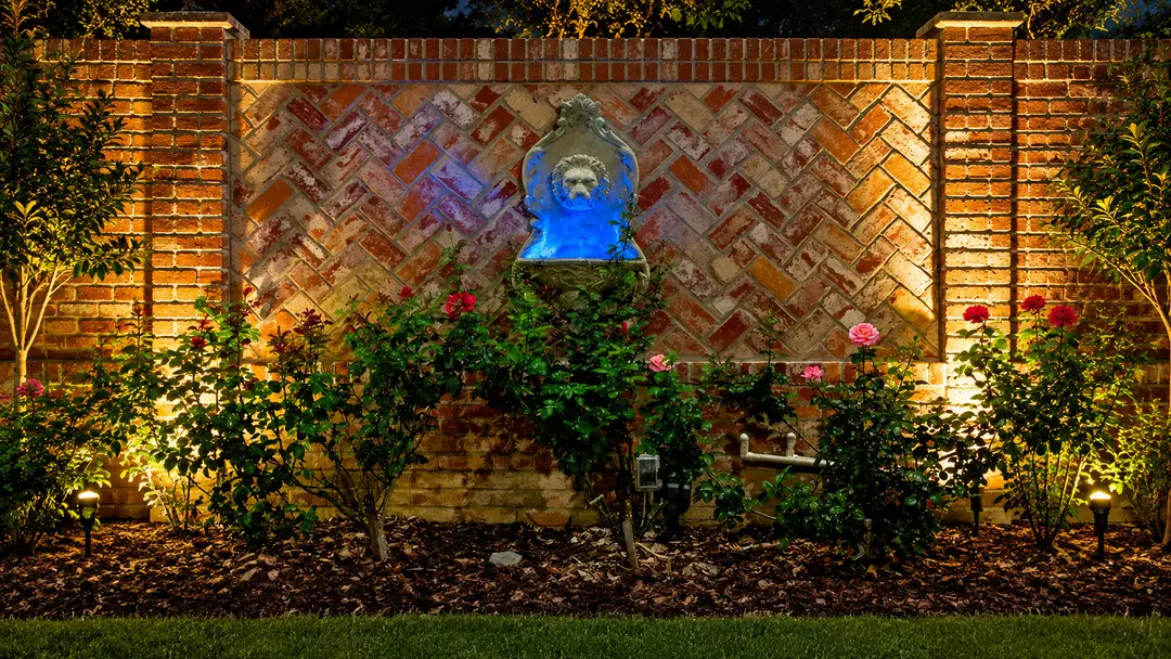 Warm landscape lighting highlights a brick courtyard wall, stone lion-head water feature, shrubs, and flowering rose bushes at night.